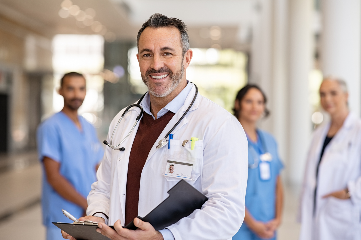 1346124955 Portrait of handsome mature doctor wearing labcoat and stethoscope reading patient case file in hospital corridor. Successful head physician standing in hospital hallway and looking at camera with his medical team in background. Reliable general practitioner in career with his healthcare staff.